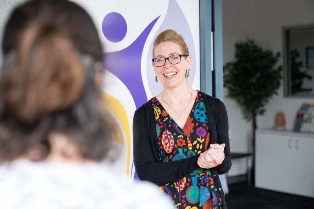 One women welcoming another into CADA. She is smiling, wearing a colourful dress with her hair pulled back. Her hands are held together, positioned like she is ready to help.