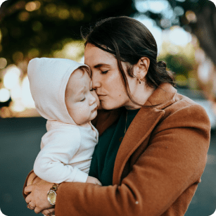 Mother holding her toddler with their faces touching and eyes closed. Mother is wearing a brown jacket, she has dark hair pulled back and light skin. Toddler is wearing a white jacket with a hood and has light skin.