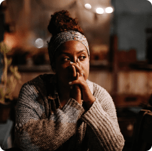 Pensive woman sitting and reflecting with hands covering mouth. Woman is wearing a light brown jumper and multi-coloured headband, she has dark brown hair tied in a bun and dark skin.