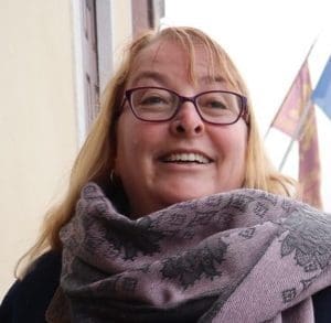 A portrait of a woman with blonde hair and glasses smiling. She is wearing a purple scarf.